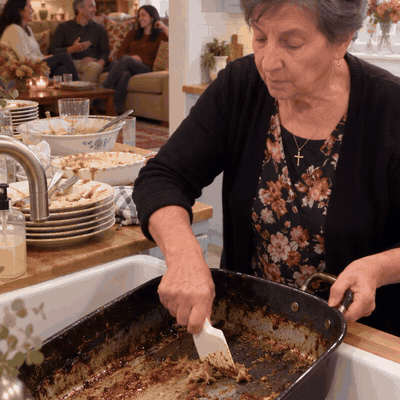 Grandma scraping a roasting pan at the sink while the family relaxes